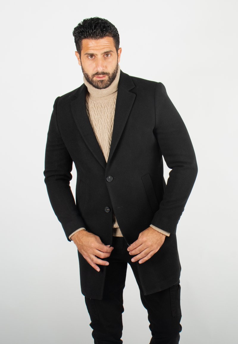 Manteau noir cintré avec deux boutons à l'avant, revers crantés et poches latérales, superposé à un pull en tricot beige à mailles torsadées, porté avec un pantalon noir.