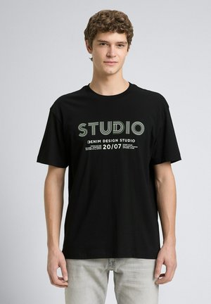Junger Mann mit lockigem Haar, der ein schwarzes T-Shirt mit dem Text „STUDIO Denim Design Studio 20/07“ trägt, steht vor einem schlichten hellen Hintergrund.