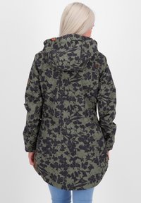 Blonde vrouw in een olijfgroene en zwarte bloemenpatroon capuchonjack, in beeld van achteren, gecombineerd met blauwe jeans.