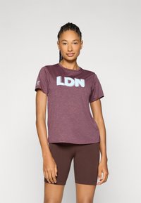 T-shirt atlético de cor bordô com "LDN" em azul claro, mangas curtas, decote redondo e tecido que afasta a umidade. Combinado com calções castanhos.