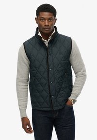 Quiltet mørkegrøn vest med brune detaljer, trykknapper og to sidelommer. Båret over en lys grå sweater, sammen med mørke jeans.
