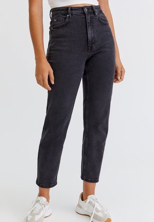 Woman wearing high-waisted black jeans and white sneakers, standing against a plain light gray background.