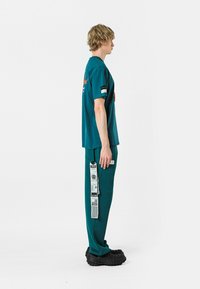 Camiseta de gran tamaño en color azul verdoso, de manga corta, con detalles impresos. Pantalones azul verdoso con bolsillo lateral, etiqueta gráfica y calzado negro texturizado. Vista lateral.