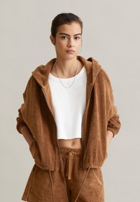 Veste à capuche zippée marron en tissu éponge doux, associée à un crop top blanc à côtes et à un short marron assorti avec des cordons de serrage.