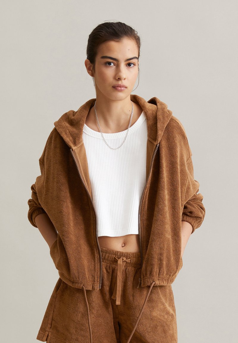 Veste à capuche zippée marron en tissu éponge doux, associée à un crop top blanc à côtes et à un short marron assorti avec des cordons de serrage.