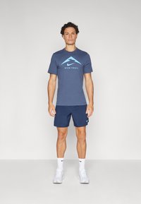 Nike Trail spordirõivad, mis sisaldavad tumesinist lühikeste varrukatega särki helebeeži mäe logoga, koos tumesiniste lühikesed pükste ja valgete tossudega.