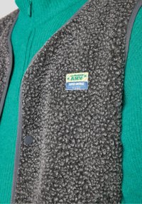 Grau Fleece-Weste mit strukturierter Oberfläche, ausgestattet mit einem Patchlabel und grauem Besatz. Trägt man über einem grünen Reißverschluss-Oberteil.