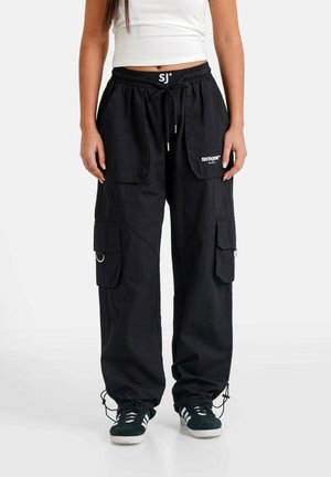 Frau trägt schwarze Cargohosen mit mehreren Taschen und schwarze Sneakers, kombiniert mit einem weißen kurzen Tanktop, stehend auf einem weißen Boden.