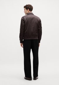 Veste bombardier marron en tissu lisse et léger, avec un ourlet et un col côtelés. Portée avec un pantalon noir légèrement fuselé.