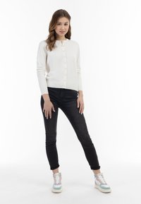 Weißer Langarm-Cardigan mit Knöpfen, kombiniert mit schwarzen Skinny-Jeans und mehrfarbigen Sneakers mit pastellfarbenen Akzenten und Schnürsenkeln.