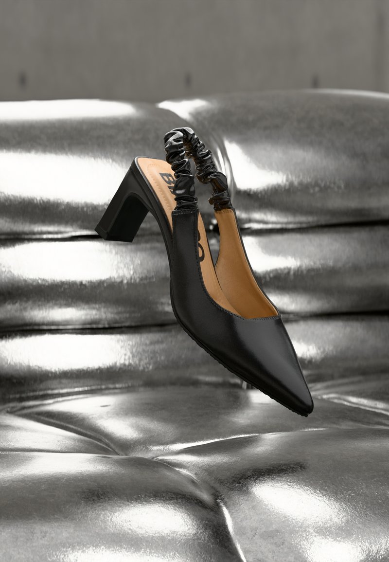 Μαύρο ψηλοτάκουνο με λουράκι στην πλάτη και μυτερή μύτη, με μαύρο ρυτιδωτό επάνω μέρος και μεσαίο χοντρό τακούνι. Λείο υλικό σε λαμπερό μεταλλικό φόντο.