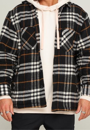 Mężczyzna w czarno-biało-brązowej kratowanej kurtce zapinanej na guziki, na jasnobeżowej bluzie z kapturem i sznurkami oraz czarnych spodniach. Pierścionki na lewej ręce.