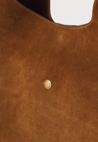 Bruine suède handtas met een gladde textuur en een goudkleurig cirkelvormig logo waarin het woord "COACH" is gedrukt.