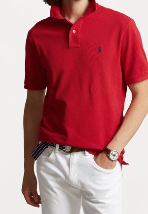 Mann trägt ein rotes kurzärmeliges Poloshirt mit gesticktem Logo, weiße Hose, gestreifter Gürtel und eine Armbanduhr, Hände in den Taschen.