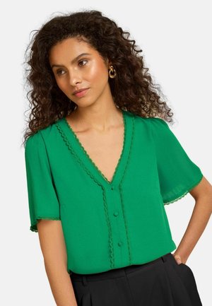 Femme aux cheveux bouclés foncés portant des boucles d’oreilles anneaux dorés et un chemisier vert à manches courtes avec bords festonnés et boutons, associé à un pantalon noir.