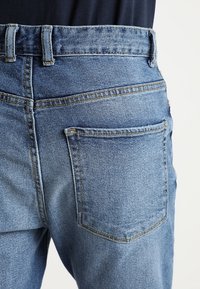 Vaqueros de mezclilla azul con una pretina alta, costuras visibles y un bolsillo trasero. La tela tiene una textura ligeramente descolorida.