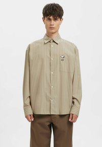 Stripete beige skjorte med knappelukking foran, lange ermer, og et tegneseriefigurmotiv på venstre brystlomme.