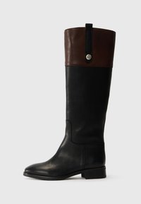 Bottines noires en cuir montant jusqu'au genou avec un revers en cuir marron, des accents en métal et une texture lisse. Dotées d'un bout rond et d'un talon plat.