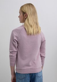 Sudadera de color rosa claro con mangas largas, cuello redondo y puños acanalados. Textura suave y ajuste relajado, combinada con vaqueros azules de mezclilla.