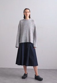 Un pull en tricot gris avec de larges manches, associé à une jupe bleu marine jusqu'aux genoux. Des sabots noirs complètent l'ensemble. Design simple et moderne.