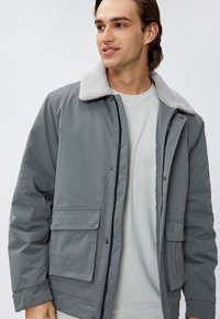 Veste grise avec un col en shearling contrastant, fermeture zip à l'avant et deux poches avant. Conçue pour un ajustement décontracté et une texture chaude.