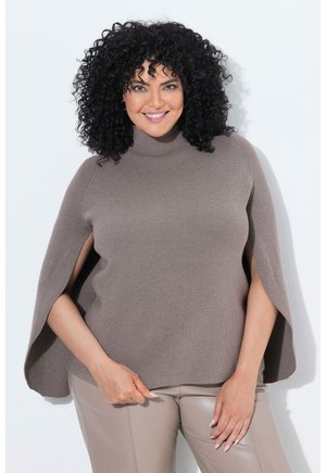 Smiling woman with curly black hair wearing a taupe high-neck cape sweater and beige pants, standing against a white background.