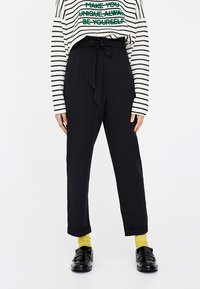 Pantalon noir taille haute avec taille nouée, jambes fuselées et texture lisse, associé à un haut à manches longues rayé blanc et des chaussettes jaunes.