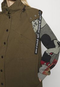 Gros plan d'une personne portant un gilet vert olive par-dessus un sweat-shirt gris avec un personnage de dessin animé rouge et noir et un ruban de texte noir sur l'épaule.