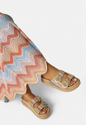 Pies con sandalias tipo slip-on entretejidas multicolores con adornos decorativos, combinadas con una falda de patrón de zigzag en tonos pastel.