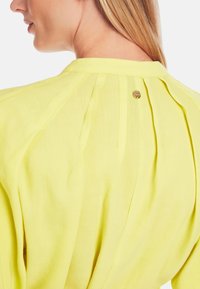 Helle gelbe Bluse mit Falten auf der Rückseite und einem kleinen goldenen Knopfdetail am Ausschnitt. Weicher, leichter Stoff mit glatter Textur.