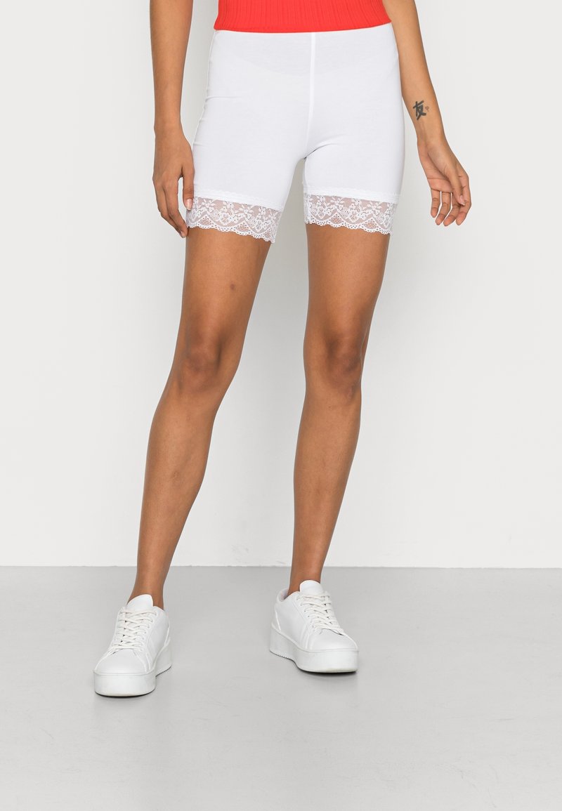 Vita stretchshorts med spetskant i fållen, kombinerade med vita sneakers. Designen är figurnära med en slät yta och en längd till mitten av låret.