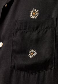 Svart stoff med en lomme som har to små trykte blomsterdesign med hvite og gule tusenfryd, og hvite knapper på venstre side.