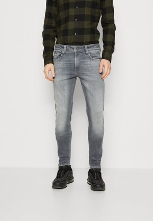 Homme portant un jean slim gris délavé, des baskets noires et une chemise à manches longues à carreaux vert foncé et noir, debout devant un fond uni.