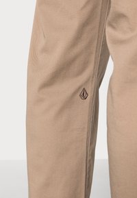 Pantalon en tissu beige présentant une texture lisse, une coupe droite et un logo brodé près de la couture latérale.