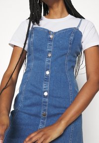 Robe en denim à coupe ajustée, de couleur bleue, avec une fermeture à boutons sur le devant et des bretelles ajustables, superposée à un T-shirt blanc.