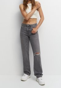 Jeans gris taille haute avec une coupe ample, présentant un détail déchiré au genou droit et un tissu en denim doux et texturé. Associés à des baskets blanches.