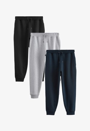 Junger Mann steht und trägt ein weißes kurzärmeliges Hemd, graue Jogginghose und schwarze Sneakers vor einem einfarbigen beigen Hintergrund.