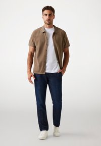 Chemise à manches courtes, en tissu texturé marron avec col, portée sur un t-shirt blanc, associée à un jean bleu foncé et des baskets blanches. Tenue simple et décontractée.