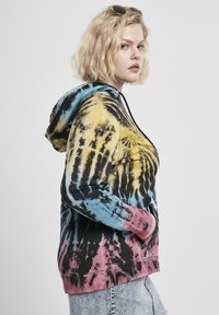 Mikina s kapucí se vzorem tie-dye v černé, žluté, modré a růžové barvě, s klokaní kapsou a stahovací kapucí. Měkká textura.