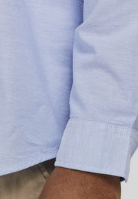 Camicia azzurra a maniche lunghe con polsini a bottone, realizzata in tessuto tessuto. Dettagli delle cuciture visibili e texture liscia sul tessuto.