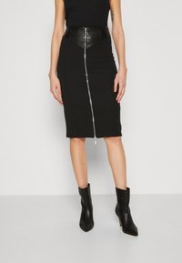 Falda lápiz negra con cintura de cuero y cremallera frontal. Diseño ajustado hasta la rodilla, combinada con botines negros de tacón.