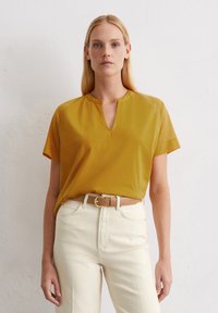 Kurzärmliges, senfgelbes Oberteil mit Stehkragen und lockerer Passform, kombiniert mit cremafarbenen Jeans und einem hellbraunen Gürtel.