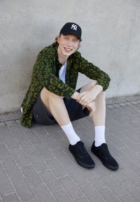 Zielona koszula w cętki, biały T-shirt, ciemne szorty, czarne sneakersy i białe skarpetki. Siedzi na utwardzonej powierzchni, oparty o ścianę.