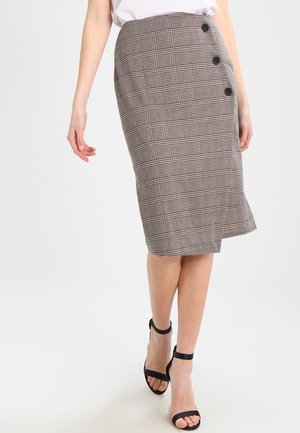 Femme portant une jupe écossaise marron longueur genou avec trois boutons noirs sur le côté, associée à des escarpins noirs à bride à la cheville.