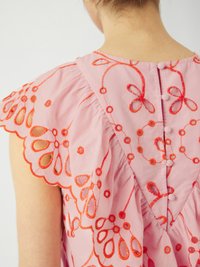Abito rosa con motivi floreali ricamati rossi, maniche arricciate e chiusura con bottoni sul retro. Tessuto di cotone morbido con bordi ondulati.