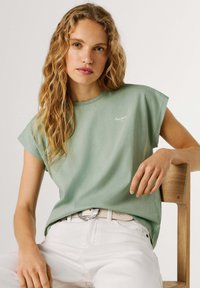 Svetlozelené bavlnené tričko s krátkym rukávom, na hrudi malým bielym logom, v kombinácii s bielymi nohavicami a ozdobným opaskom.