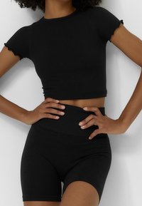 Czarny prążkowany crop top z krótkimi falbankami, zestawiony z czarnymi szortami o wysokim stanie. Miękka tekstura, dopasowany krój, bezszwowe wykończenie.