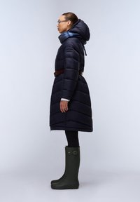 Giacca imbottita blu navy con vita cintava, dotata di cappuccio e sciarpa. Modello che indossa leggings neri e stivali impermeabili verdi.