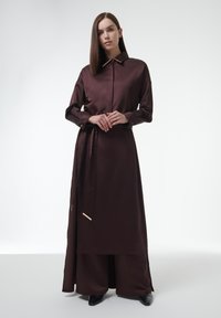 Bruine lange jurk met kraag, ceintuur in de taille en gouden hardware accenten. Gladde stof en lange mouwen, gecombineerd met brede broeken.