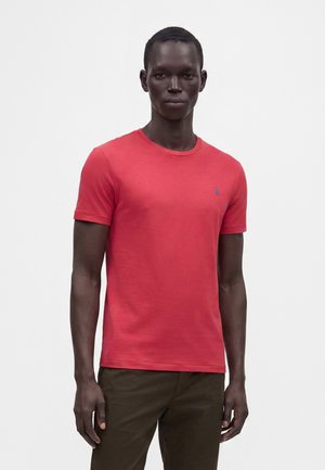 Homme portant un t-shirt rouge uni à manches courtes et un pantalon marron foncé, debout devant un fond blanc.